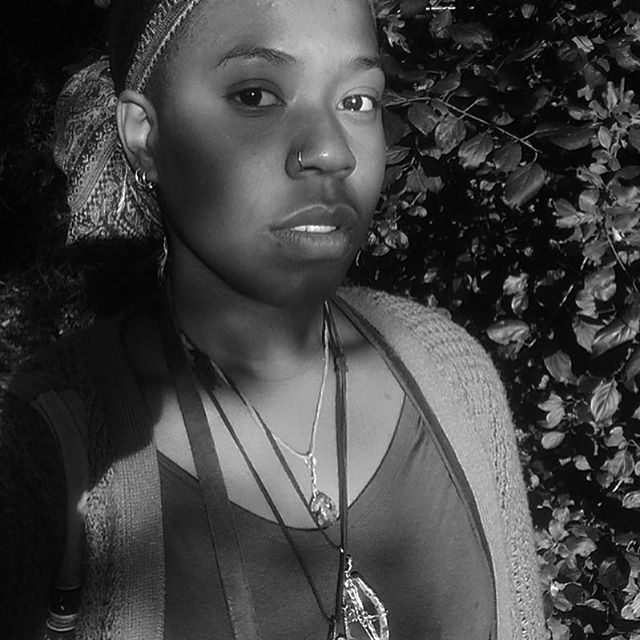 Portrait of woman with headwrap and jewelry, standing before dense foliage