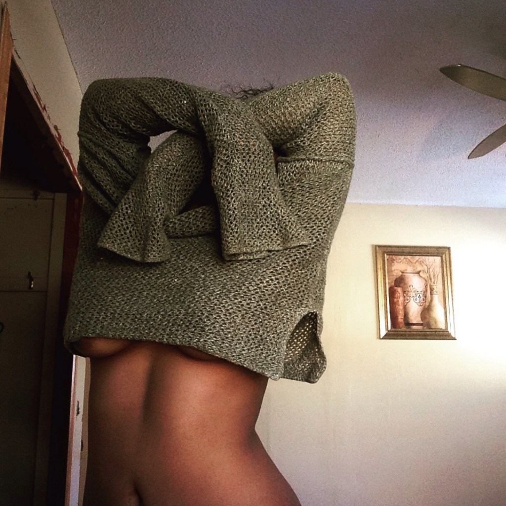 A person lifting a knit sweater over their head, revealing their bare torso, captured mid-motion in a softly lit room.