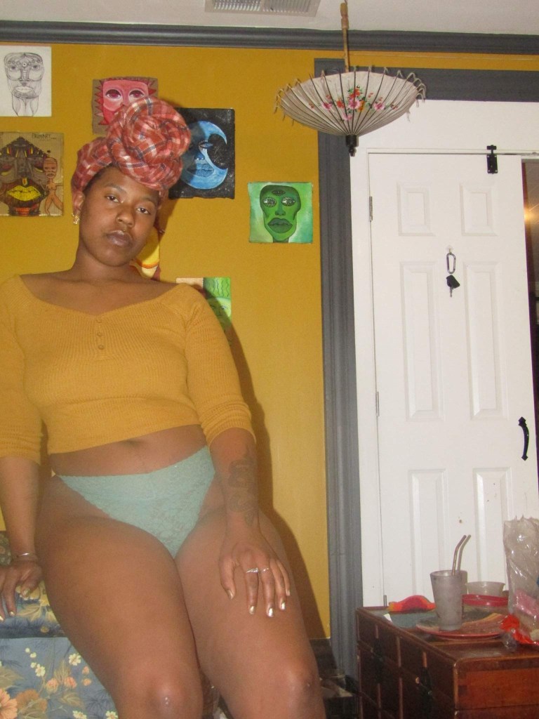 A woman sits on the edge of a bed, relaxed and poised in soft underwear and a cropped mustard top. A red headwrap crowns her, and painted portraits on a mustard yellow wall echo her gaze. Light spills in from a white door, illuminating the stillness.