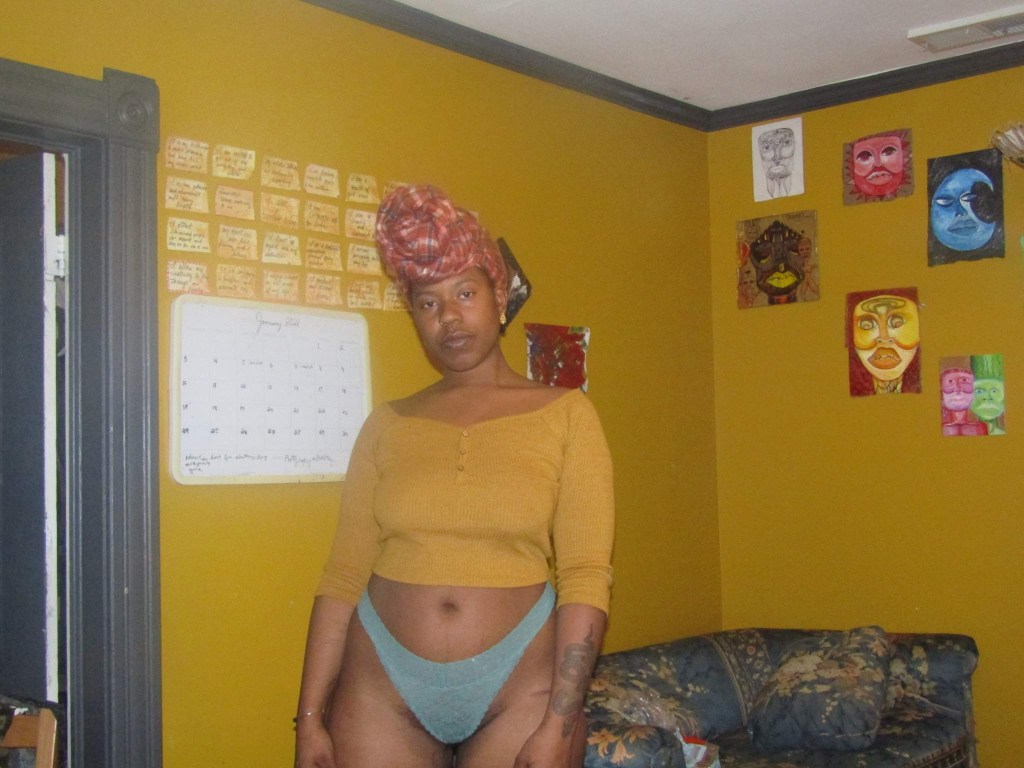 A woman stands grounded in a mustard yellow room, wearing a cropped top, underwear, and a red headwrap. Her expression is steady and unbothered, framed by a wall of colorful portraits and affirmations.