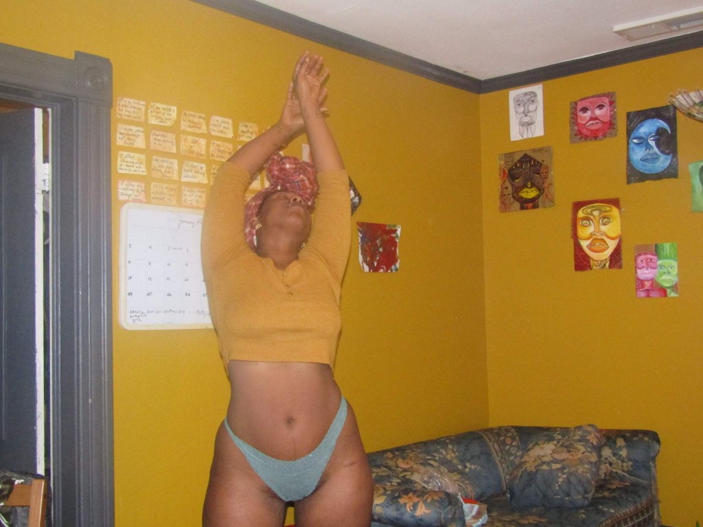 A woman stands with arms raised, palms pressed in a prayer-like gesture above her head. She wears a mustard top, green briefs, and a red headwrap. The mustard-yellow walls behind her are adorned with vivid painted faces, and a calendar hangs nearby.