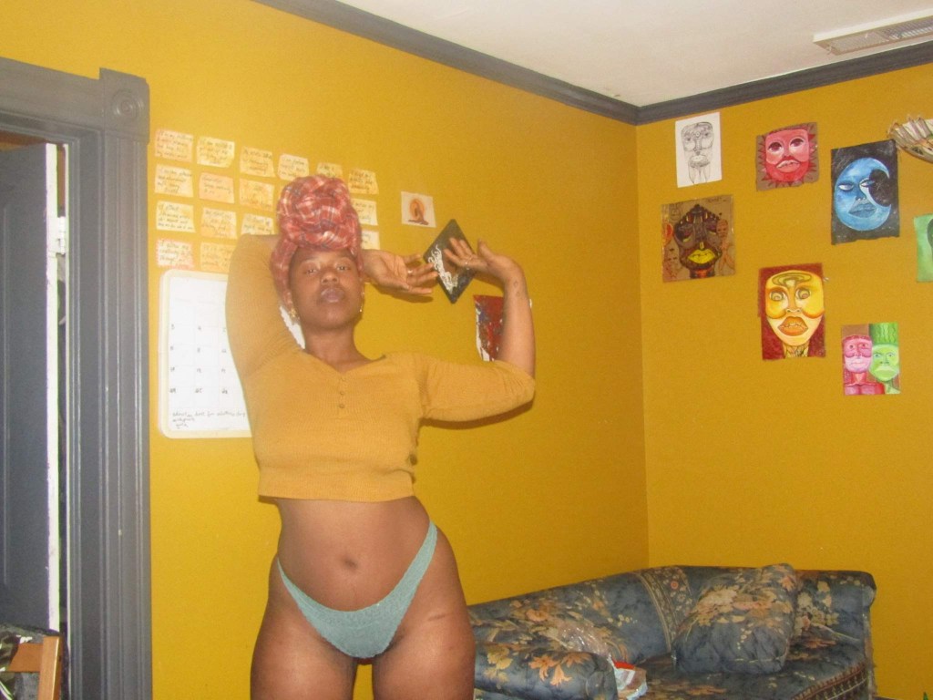 A woman stands in a mustard yellow room wearing a cropped top, underwear, and a red headwrap. Her arms are raised in a fluid, expressive gesture as if caught mid-dance or invocation. Art-covered walls and a floral couch frame the space with bold, lived-in warmth.