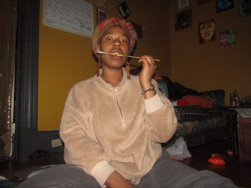 A woman sits on the floor in a cozy room, holding a paintbrush between her teeth.