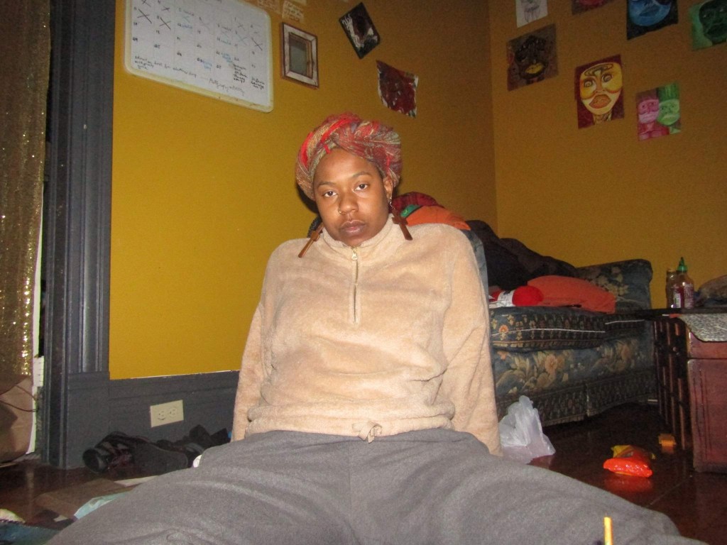 Woman sitting on the floor in a cozy tan pullover and headwrap, gazing directly at the camera in a warmly lit room.