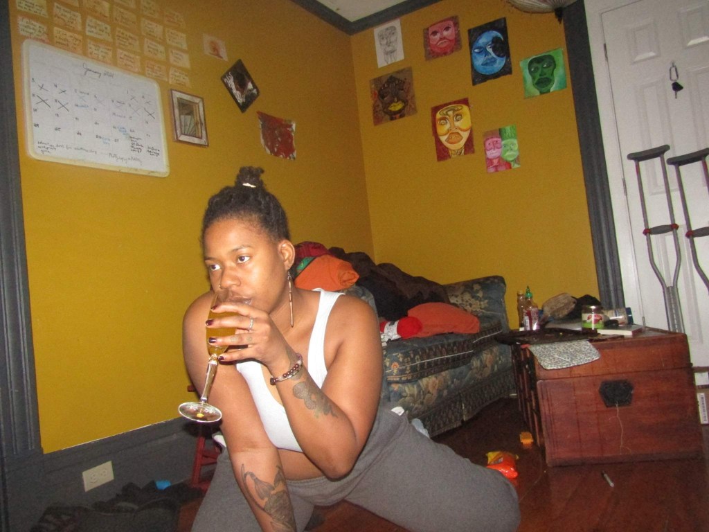 Woman in a white crop top and gray pants crouched on the floor, sipping from a champagne glass in a room with mustard yellow walls and expressive artwork.