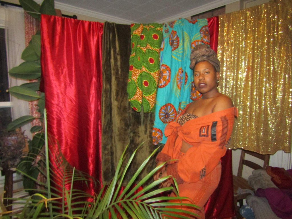 Black woman in burnt orange and leopard print standing among vibrant hanging fabrics and indoor plants.