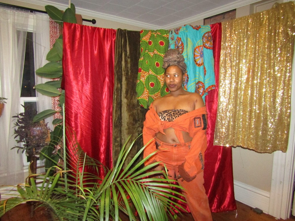 Black woman in an orange outfit standing confidently amid colorful drapes and lush indoor plants.
