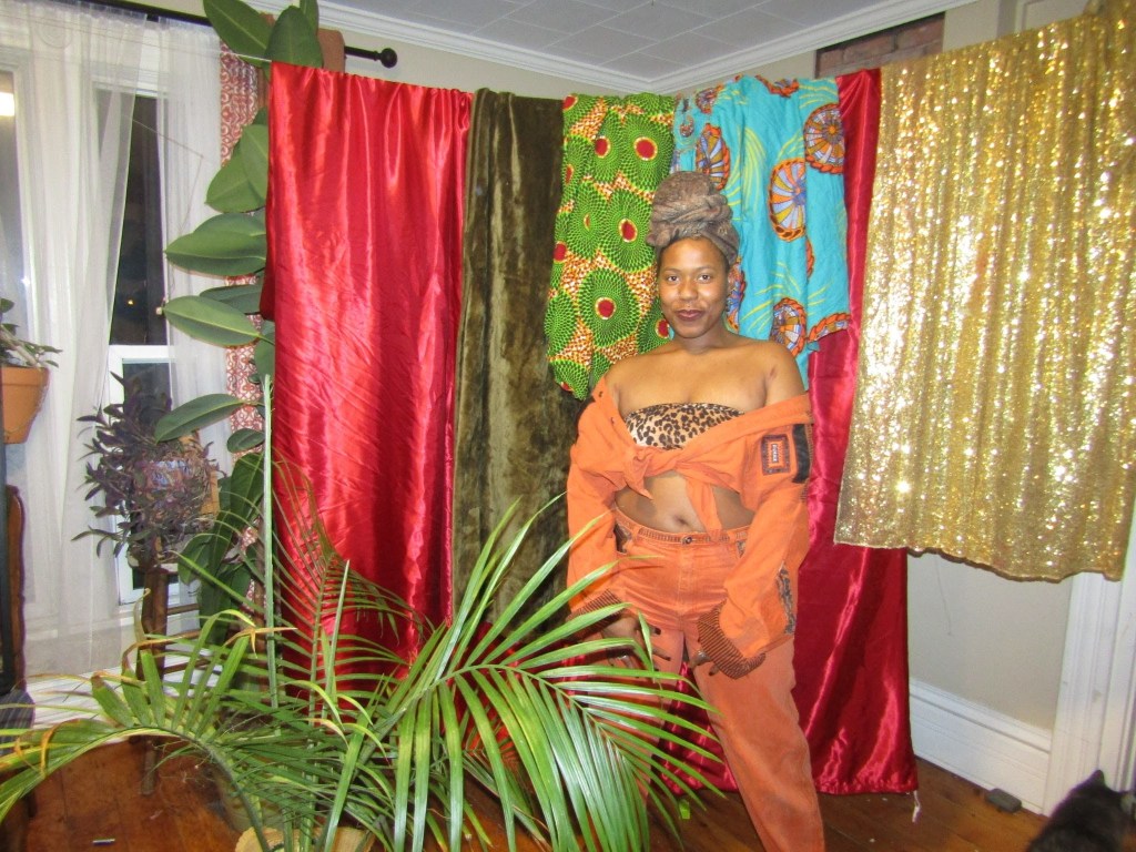 Afrocentric woman in orange two-piece and leopard bandeau, posing confidently in front of colorful draped fabrics and lush indoor plants.