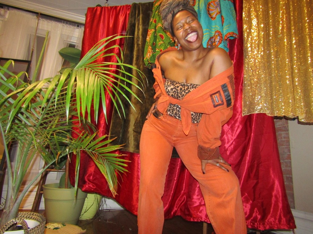 Playful woman in orange outfit and leopard bandeau posing joyfully with tongue out, surrounded by vibrant Afrocentric fabrics and lush plants.