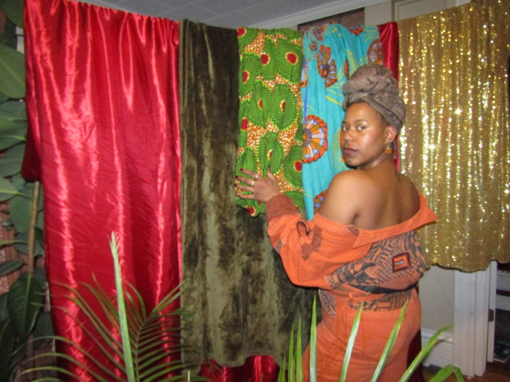 Woman in orange and brown turns to face the camera while touching vibrant African fabrics hanging beside her.