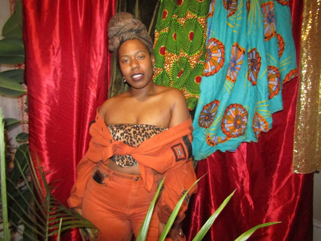 Woman in an orange two-piece and leopard bandeau standing confidently in front of vibrant Afrocentric fabric and lush plants.