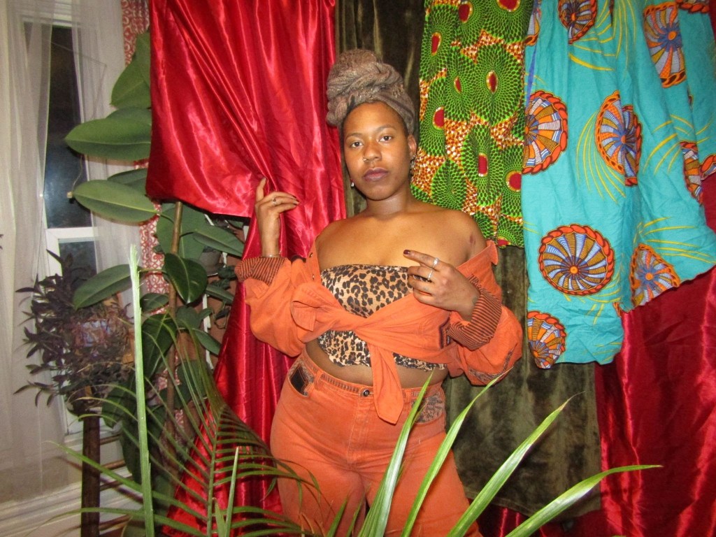 Black woman in orange two-piece and leopard bandeau standing confidently among vibrant fabrics and plants.