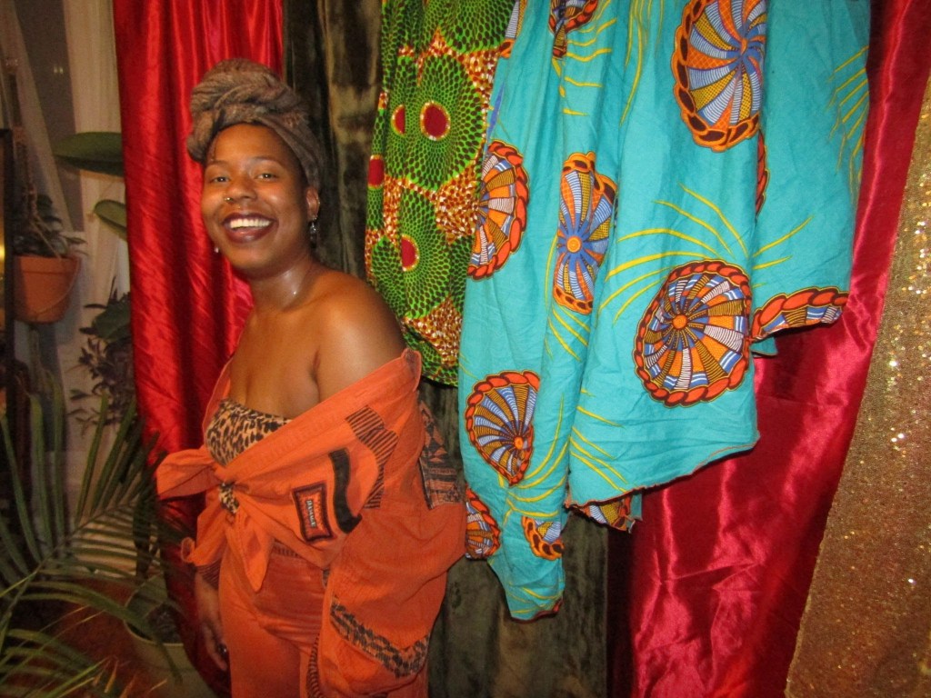 Smiling Black woman in orange outfit standing beside colorful patterned fabrics and plants.