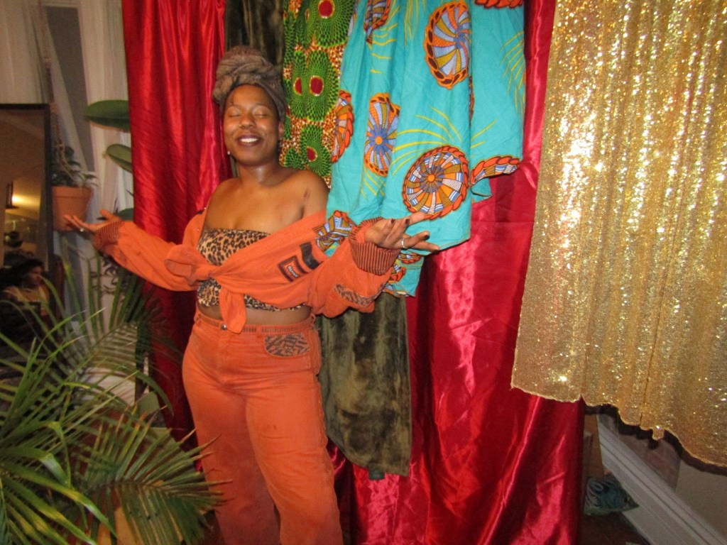 Black woman smiling joyfully in an orange outfit, surrounded by vibrant drapes and cultural fabrics.