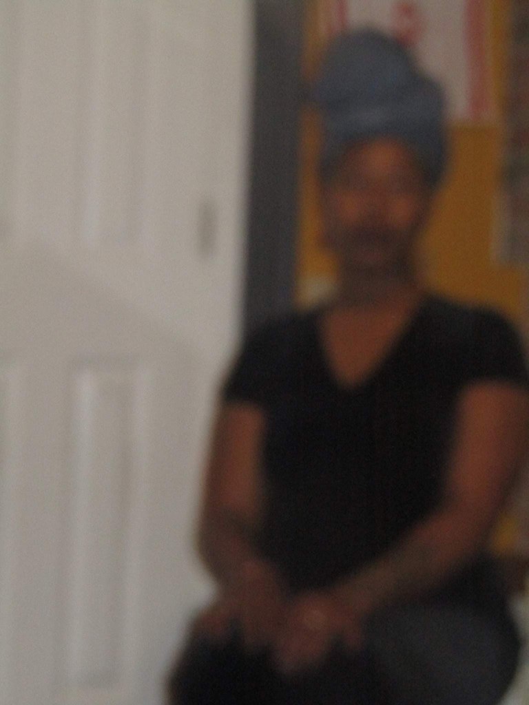 A soft, unfocused image of a woman in a black dress and blue headwrap, seated in a room with warm walls and a closed white door.
