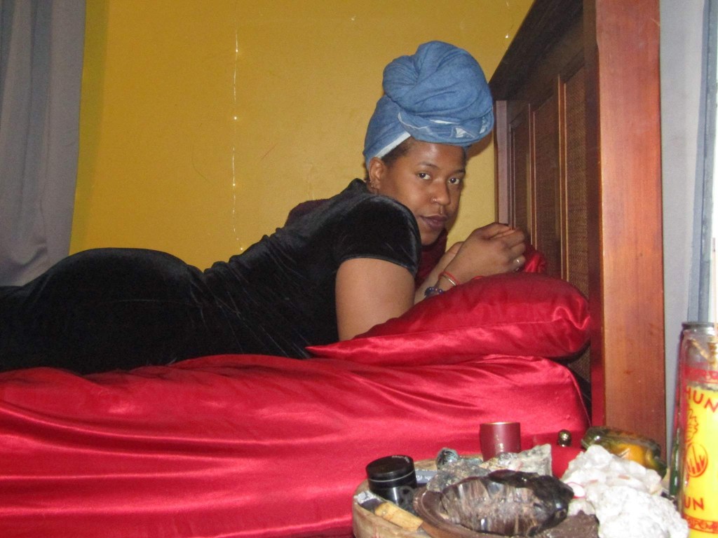 A woman lies on her stomach on a red satin bed, wearing a black velvet dress and a blue towel wrap on her head. She looks calmly into the camera. A bedside altar with candles, crystals, and sacred items rests nearby.