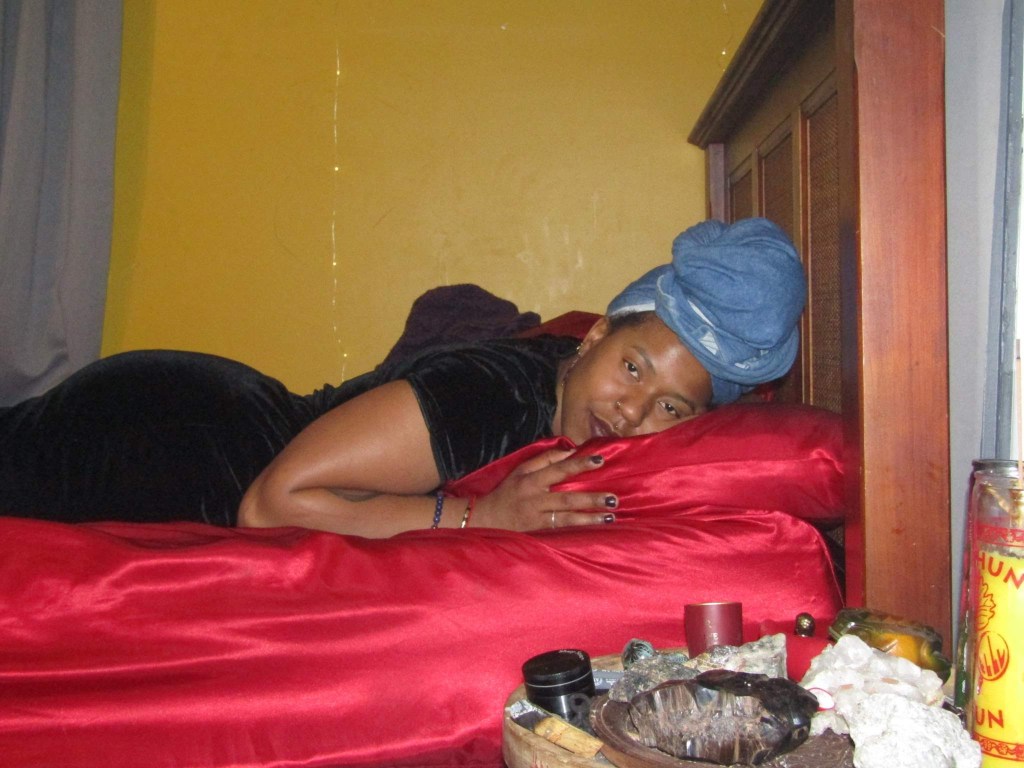 A woman lies on red satin bedding with her head resting on a matching pillow, eyes softly gazing at the camera. She's wrapped in a dark velvet dress and a blue towel, beside a wooden headboard and an altar of candles, oils, and crystals.