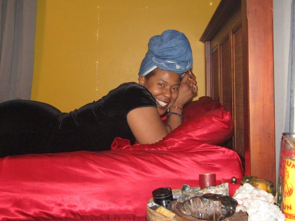 A smiling woman lies on a red satin bed, wearing a black velvet dress and a denim headwrap. Her face is joyful, framed by cozy bedroom textures and ritual items beside her.