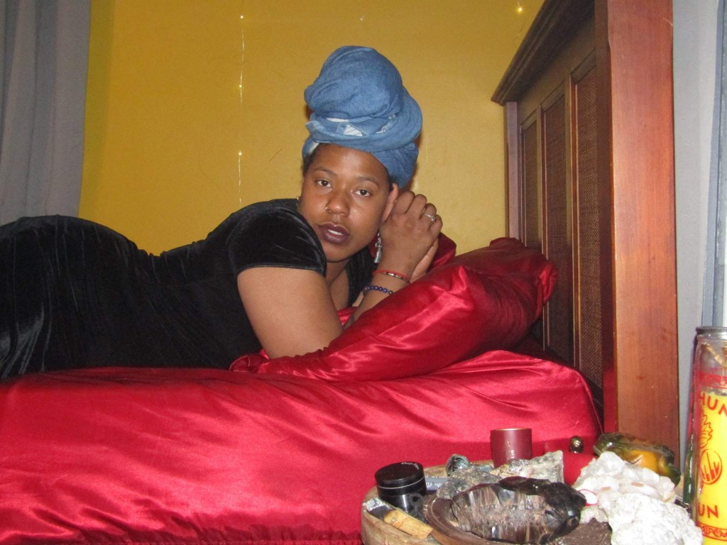 A woman reclines on a red satin bed, head turned softly toward the camera. She wears a denim headwrap and black velvet dress. A tray of candles, crystals, and sacred objects rests nearby.
