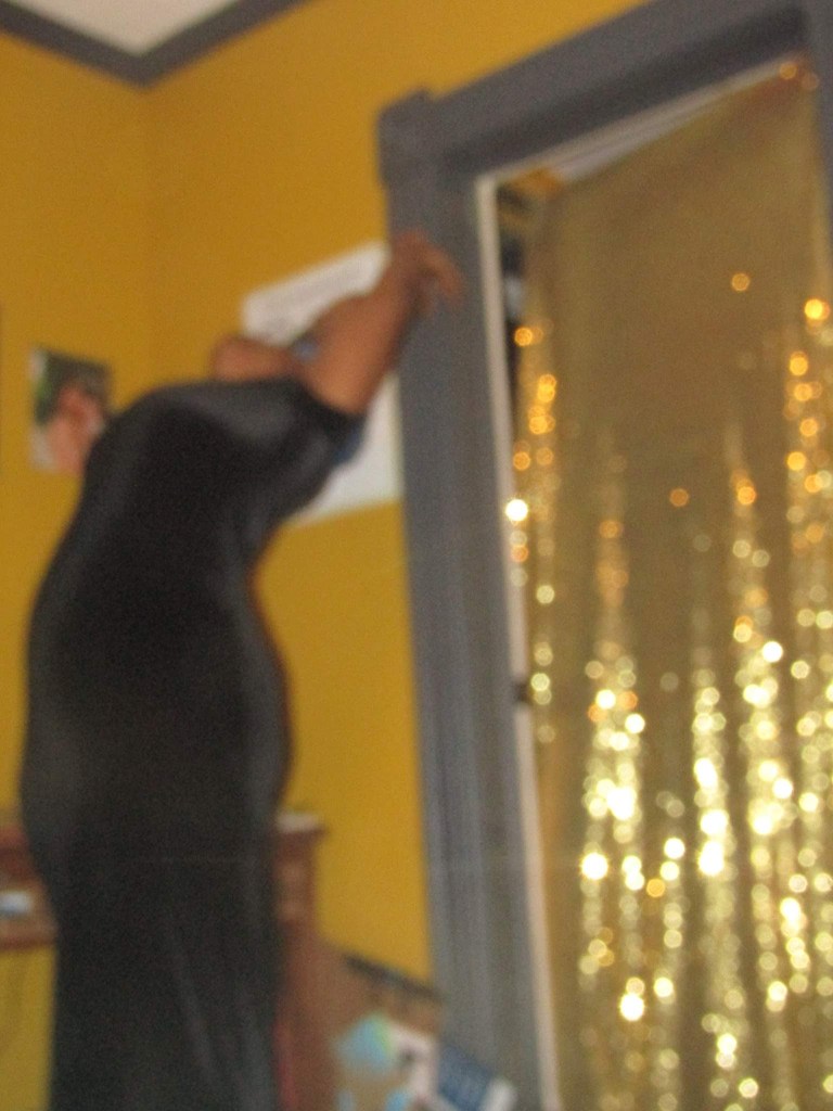 A blurred figure in a black dress stretches upward toward a golden curtain, framed by mustard-yellow walls and a dark doorway.