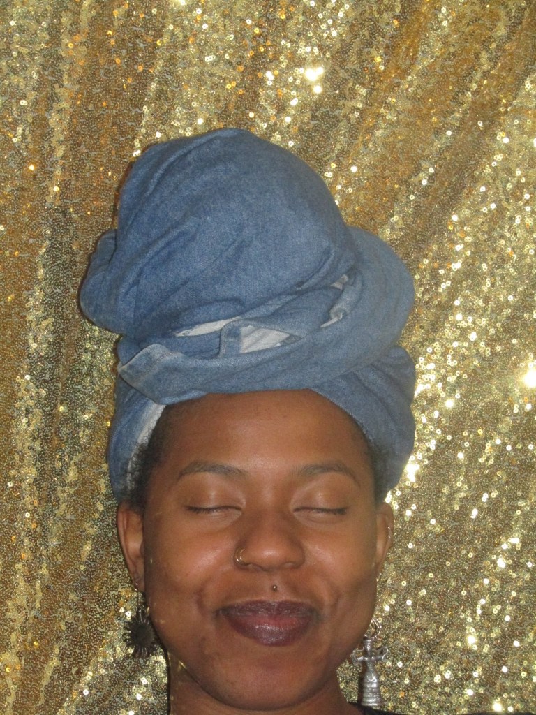 A close-up of a woman in a denim headwrap smiling gently with her eyes closed in front of a sparkling gold sequin backdrop. Her expression is serene and joyful.