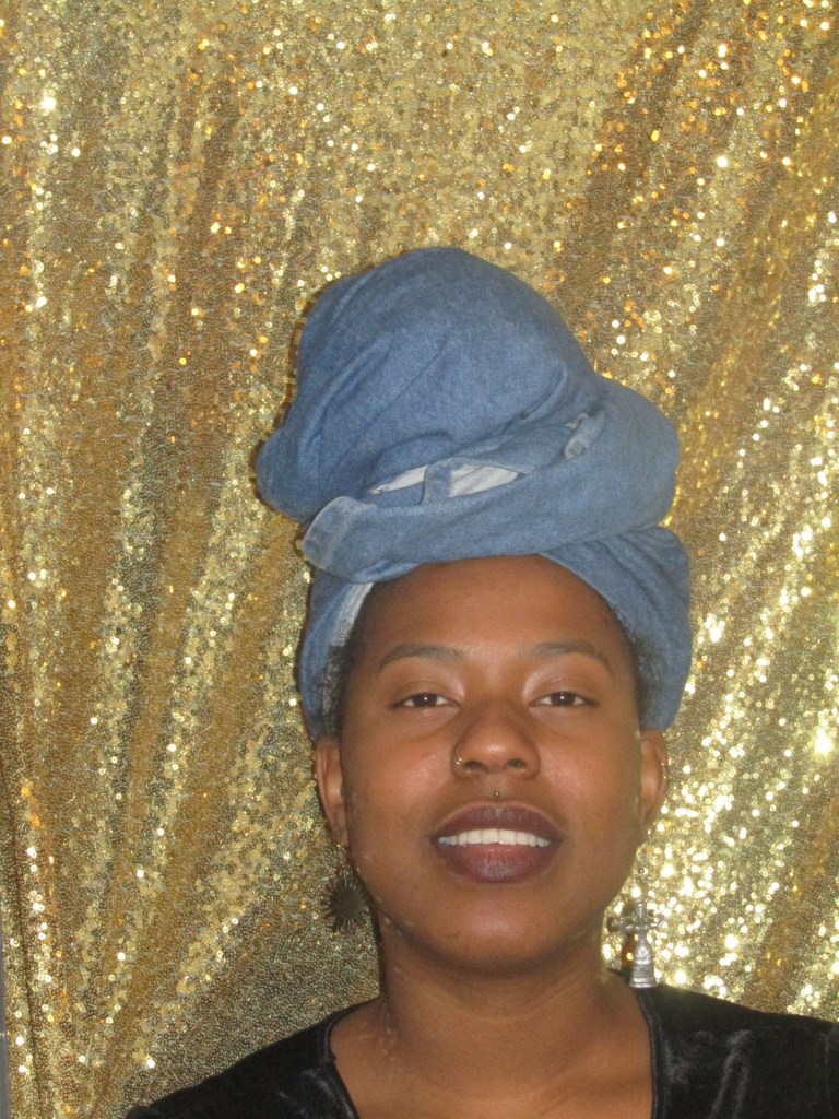 A close-up portrait of a woman wearing a denim headwrap and dark lipstick, standing before a shimmering gold sequin backdrop. She looks directly at the camera with a soft, grounded expression.