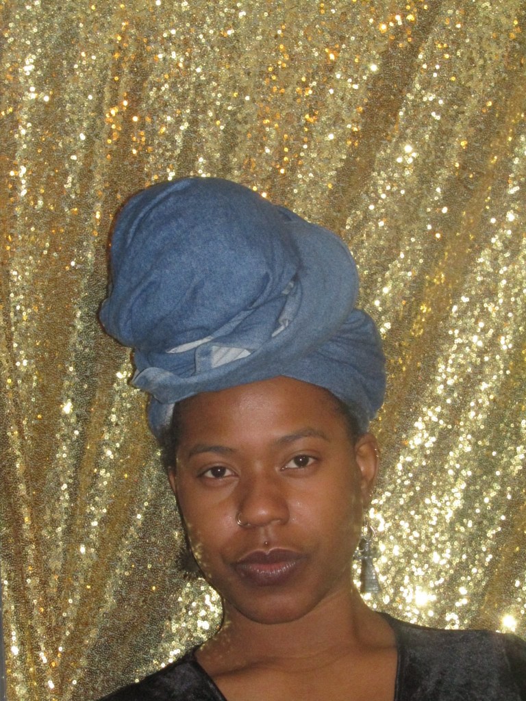 A close-up portrait of a woman in a denim headwrap, standing in front of a shimmering gold backdrop. Her expression is calm and serious, exuding quiet strength.