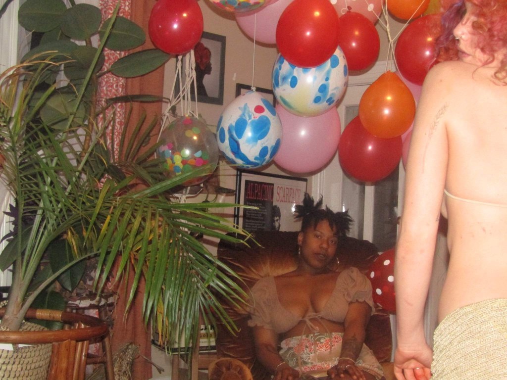 Two women in a softly lit room decorated with vibrant balloons and lush houseplants. One woman sits in a golden chair with a calm expression, while another stands in the foreground with her back turned, partially visible. The scene is warm, dreamy, and candid.