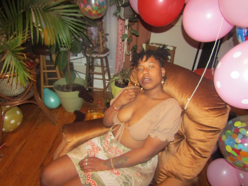 A woman reclines in a golden chair surrounded by balloons, plants, and soft lighting. She gazes upward, one hand to her chest, embodying both ease and quiet intensity.
