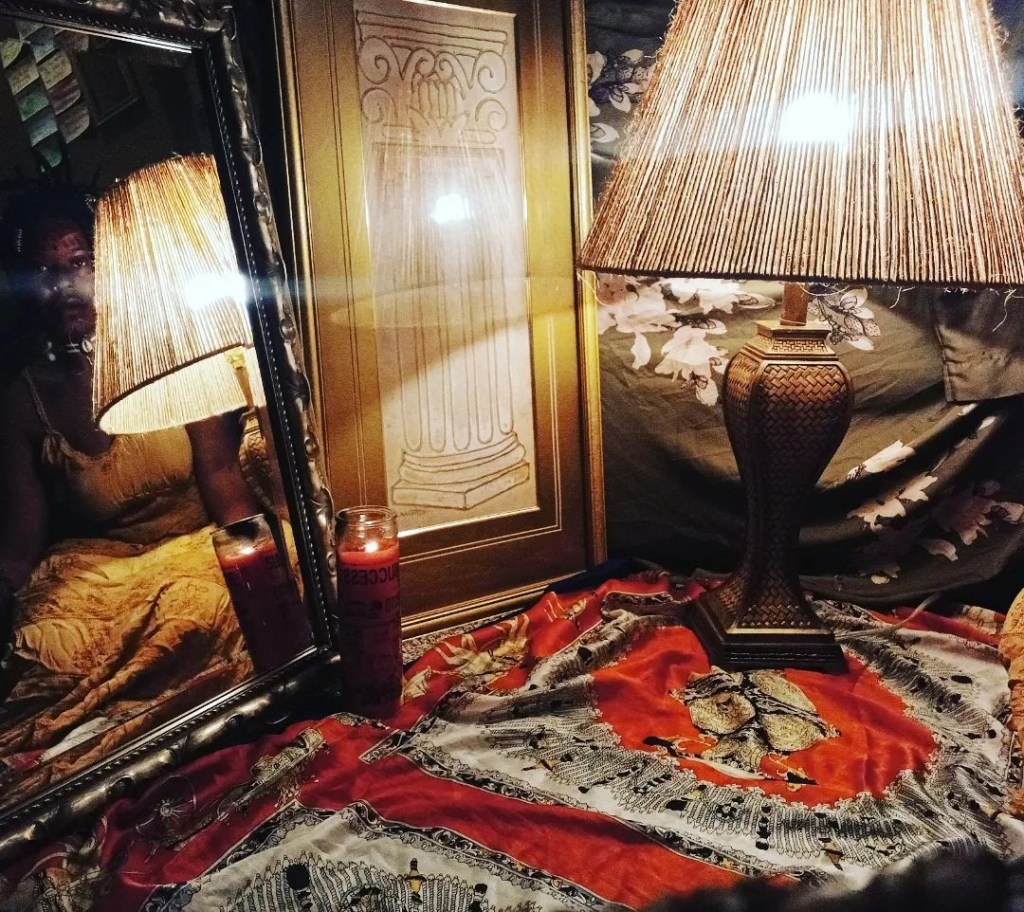 A glowing lamp sits on a richly patterned cloth beside a framed column drawing, a red candle, and a mirror reflecting part of a woman's body in a yellow dress. The setup feels sacred, intimate, and mystical, bathed in warm amber light.