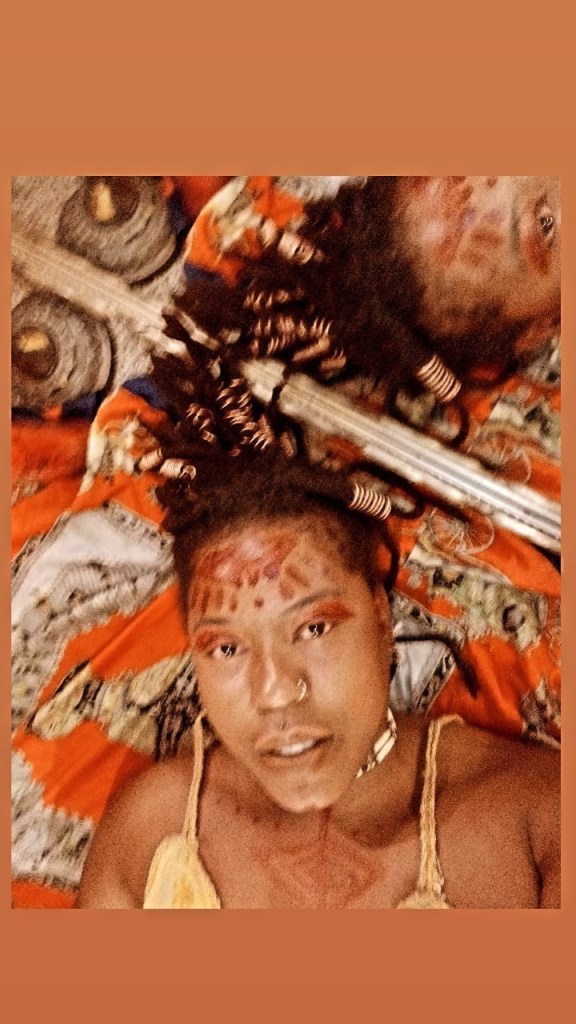 A woman lies on vivid patterned cloth with ritual markings adorning her face and chest. Her hair fans outward like a crown, partially reflected in a mirror beside her. The image radiates warmth, ancestral symbolism, and sacred femininity.