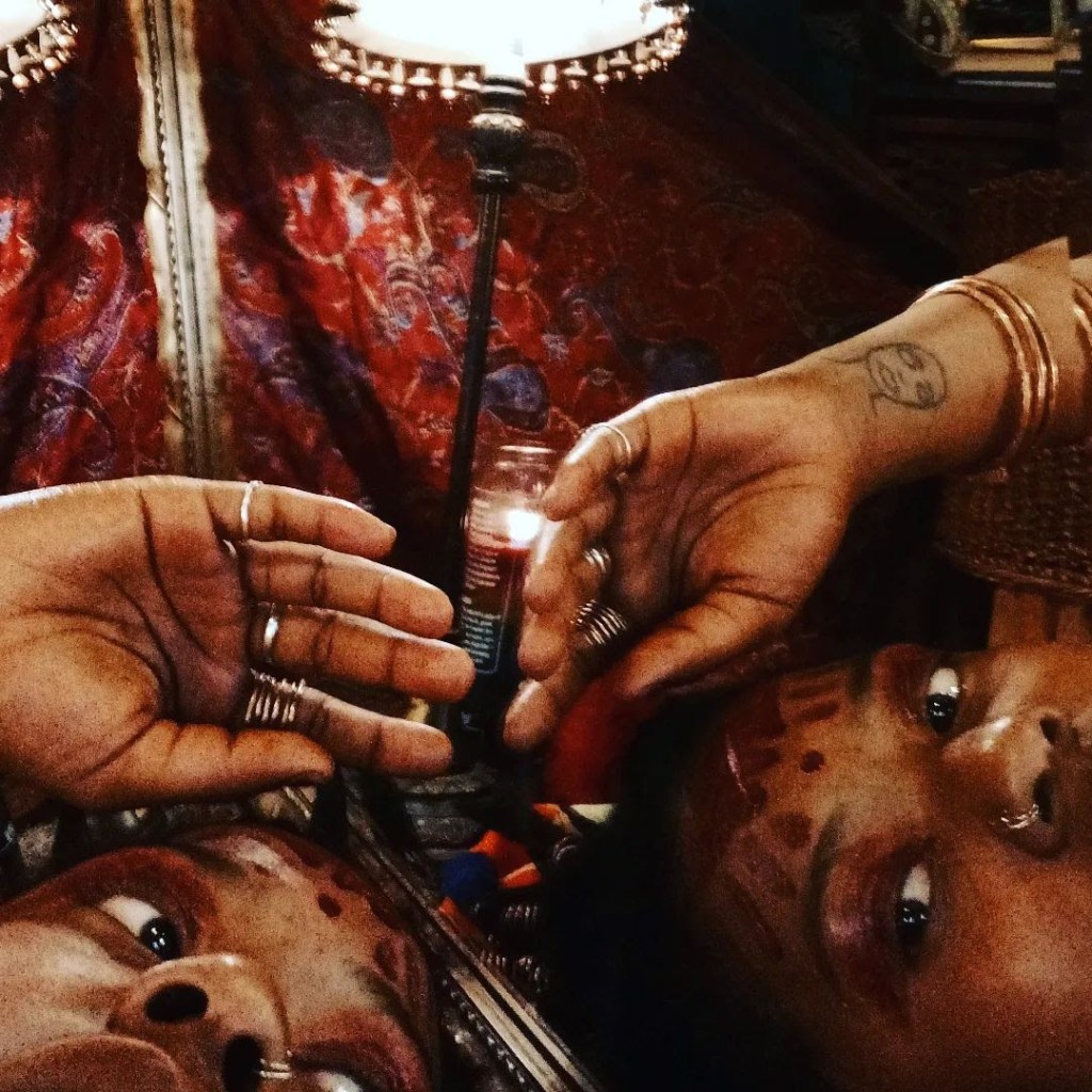 Two mirrored reflections of a woman with ritual face markings, jewelry, and henna-like tattoos. Her hand rests across the mirror with rings and bangles, under a warm lamp.