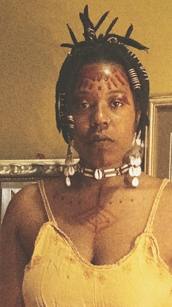 A woman stands facing the camera, adorned with shell jewelry and red ritual markings across her forehead, chest, and shoulders. Her gaze is steady and ancestral. Hair crowned with spikes and beads, she wears a yellow top and stands in front of a mirror. The atmosphere is intimate and reverent.