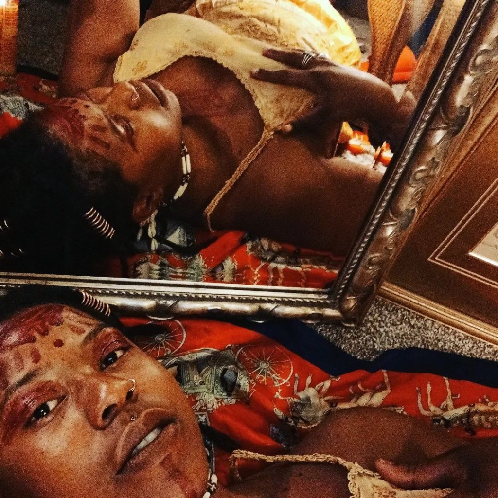 A woman with sacred markings lies beside a mirror, her reflection doubling the intimate gaze. She wears a cream dress and lies on a vivid red and blue patterned cloth, surrounded by gold frames and ritual stillness.