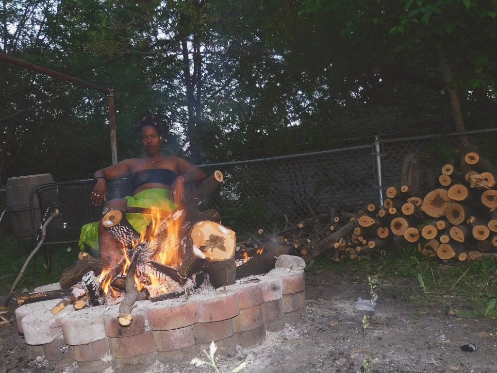 A woman in a strapless top and bright green skirt sits behind a roaring outdoor fire pit. Her posture is powerful and relaxed, framed by cut logs stacked to the side and trees overhead. She appears calm, commanding, elemental.