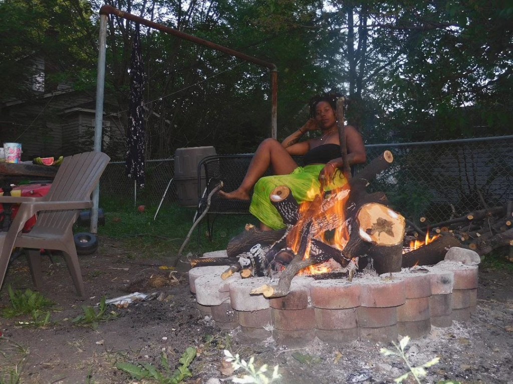 A woman in a lime green skirt sits beside a fire pit made of bricks, surrounded by chairs, logs, and soft dusk light. She reclines calmly while flames blaze in front of her, the yard dim and overgrown in the background.