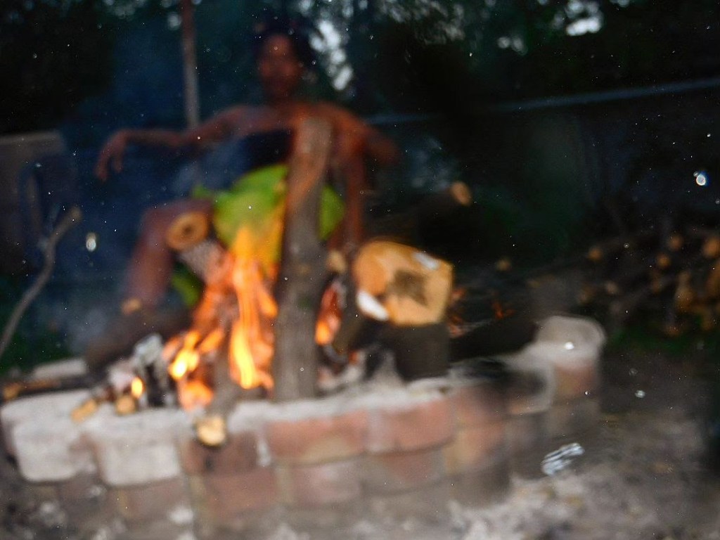 A blurred figure of a woman sits behind a fire pit, her body nearly dissolving into smoke and shadow. The flames leap in the foreground, casting light and movement into the darkening outdoor space.