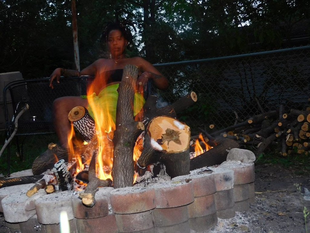 A woman sits behind a blazing outdoor fire pit, her form partially cloaked in flames and shadows. Logs burn intensely in front of her, glowing orange against the dusk. Her presence is steady, powerful, almost spectral.