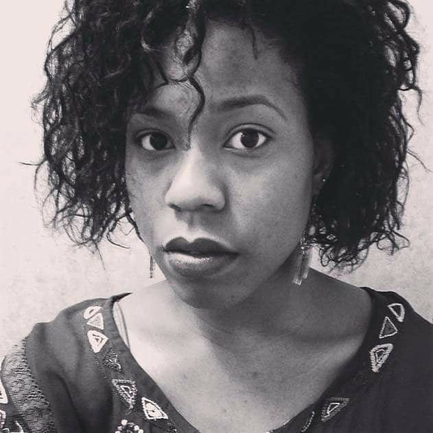 Black-and-white close-up portrait of a young woman with natural curls and geometric earrings. She gazes calmly and directly at the camera, wearing a patterned top.