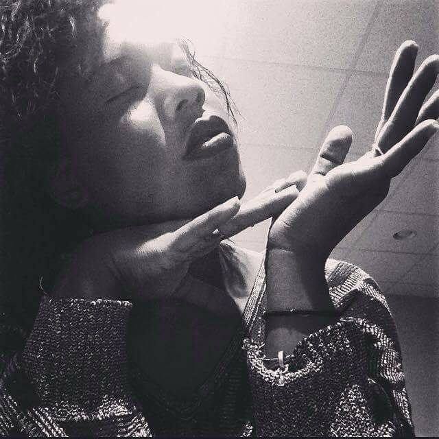 Black-and-white image of a woman with eyes closed, tilting her face upward with hands held delicately near her chin, mid-expression, under soft ceiling light.