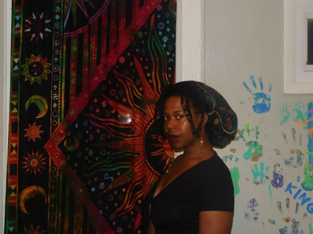 A woman in a black top stands in profile, her gaze direct and soulful. She wears a scarf wrapped over her locs, adorned with beads. Behind her, a vibrant cosmic tapestry glows with suns, moons, and stars. A wall of colorful handprints and children's scribbles surrounds her, contrasting the mystic backdrop.
