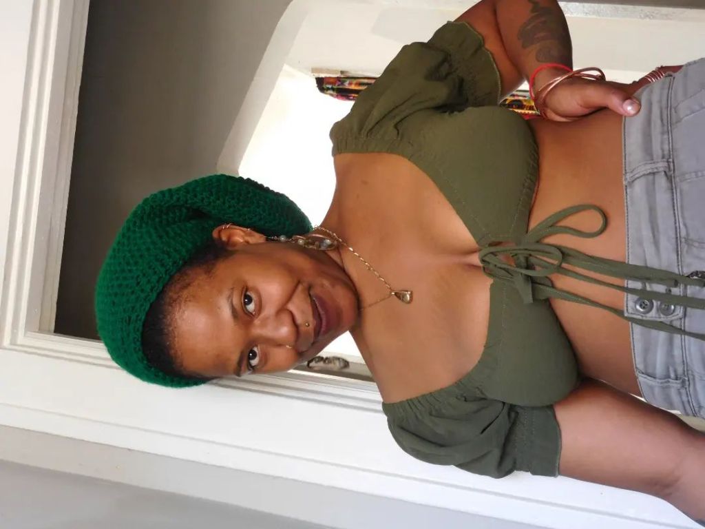 A woman stands confidently in a doorway, her body turned slightly as she gazes into the camera with a knowing smile. She wears an olive green tie-front top, denim shorts, and a green knit beret. Her jewelry is subtle yet present, and her expression holds warmth with a playful edge.