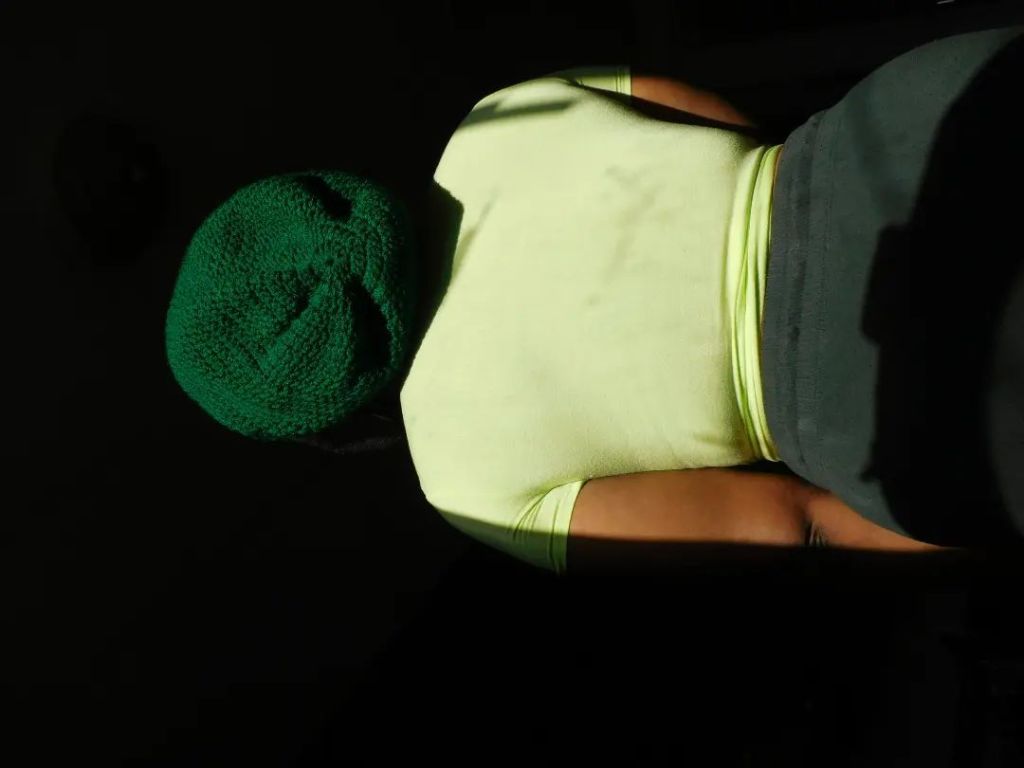 A woman stands with her back turned, bathed in golden sunlight. She wears a green crocheted hat and a pale yellow top, blending into the dark backdrop like a quiet silhouette.