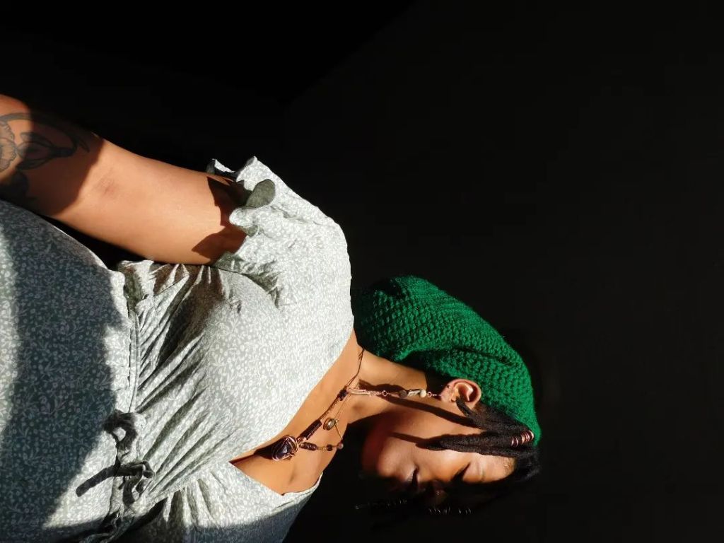 A woman in a soft sage dress and green knit beret stands against a deep black background, her face turned down in quiet grace. Braided strands fall over her cheek, and sunlight brushes her skin like a blessing.