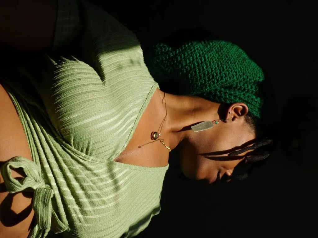 A close-up of a woman in a light green ribbed top and a forest green knit beret, her head tilted downward in soft shadow. Her lips are parted, locks fall over her cheek, and sunlight dances across her chest and collarbone.