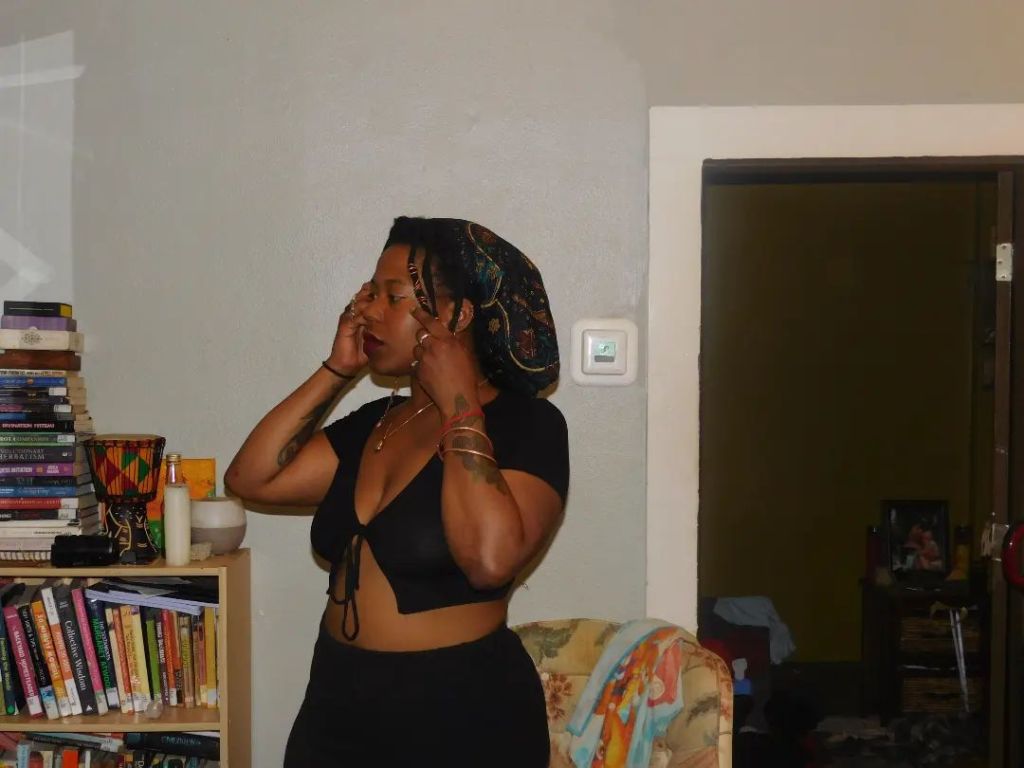 A woman in a black top adjusts her headwrap while standing near a bookshelf. Her posture is relaxed yet focused, framed by warm lighting and a cozy, book-filled room.