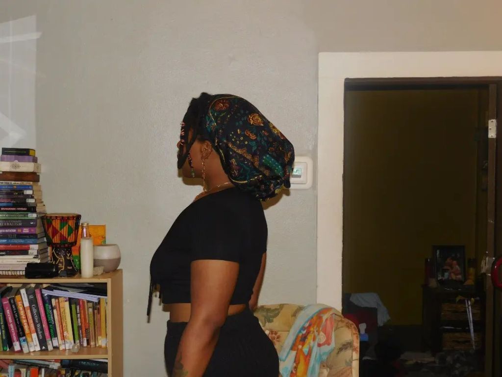 A woman stands in profile, facing a bookshelf filled with colorful titles. She wears a patterned headwrap and a black cropped top, standing in still focus within a cozy, lived-in room.