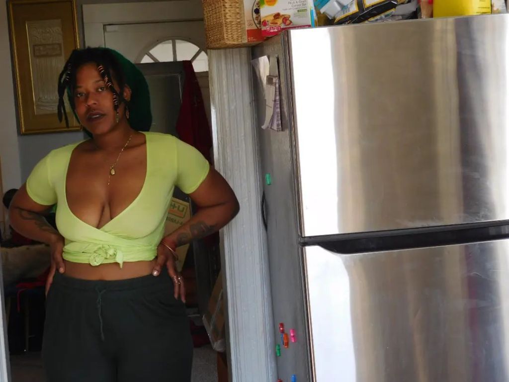 A woman in a lime green crop top and dark pants stands confidently in a kitchen doorway beside a stainless steel fridge. Her hands are on her hips, her locs are adorned with beads, and a soft but grounded gaze meets the lens.