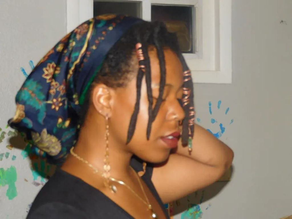 A woman in profile with locs adorned in beads and a patterned headscarf. Her eyes are closed, lips painted deep red, one hand raised behind her head. The background features teal handprints on a gray wall.