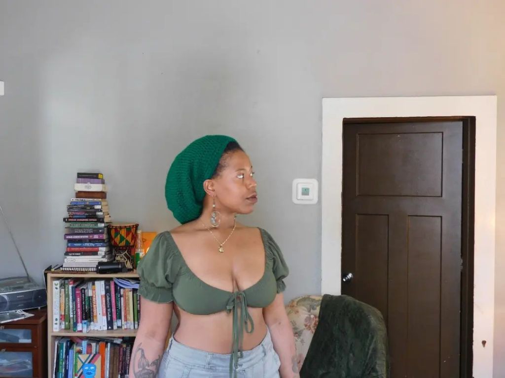 A woman stands indoors, wearing a green knit hat and matching top, with light denim jeans. Her gaze is lifted to the side as if answering an unspoken call. Behind her is a shelf stacked with books and a dark wooden door, evoking a lived-in, thoughtful space.